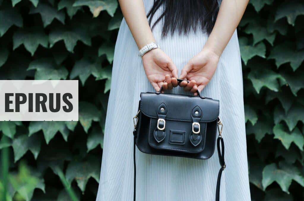 A woman in a striped dress holds a black leather handbag with lush green leaves in the background.